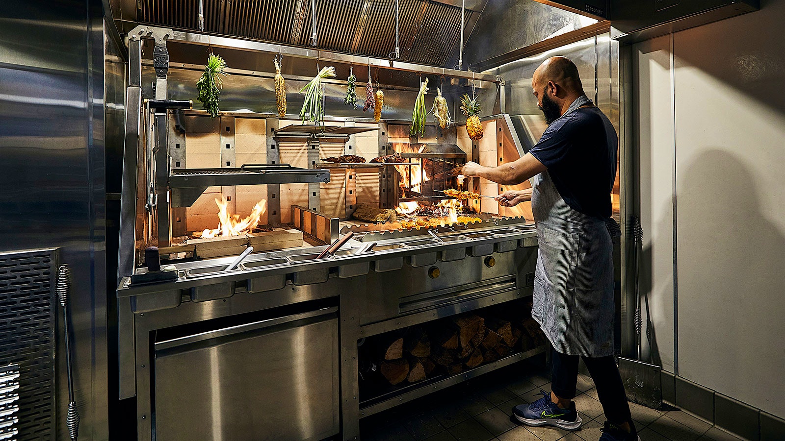 Chef Sujan Sarkar cooking at a grill, with corn and other vegetables hanging above in the kitchen, at the restaurant Indienne in Chicago